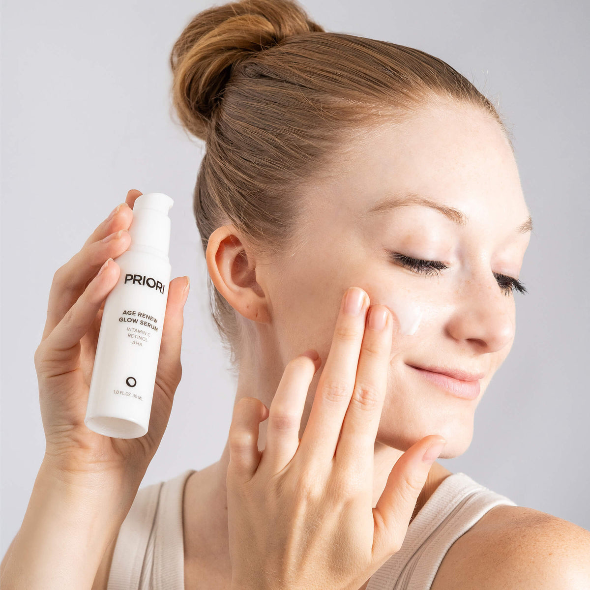 A woman applies anti aging cream from a PRIORI Age Renew Glow Serum with Retinol bottle to her cheek, holding the white pump bottle in her hand against a light gray background.