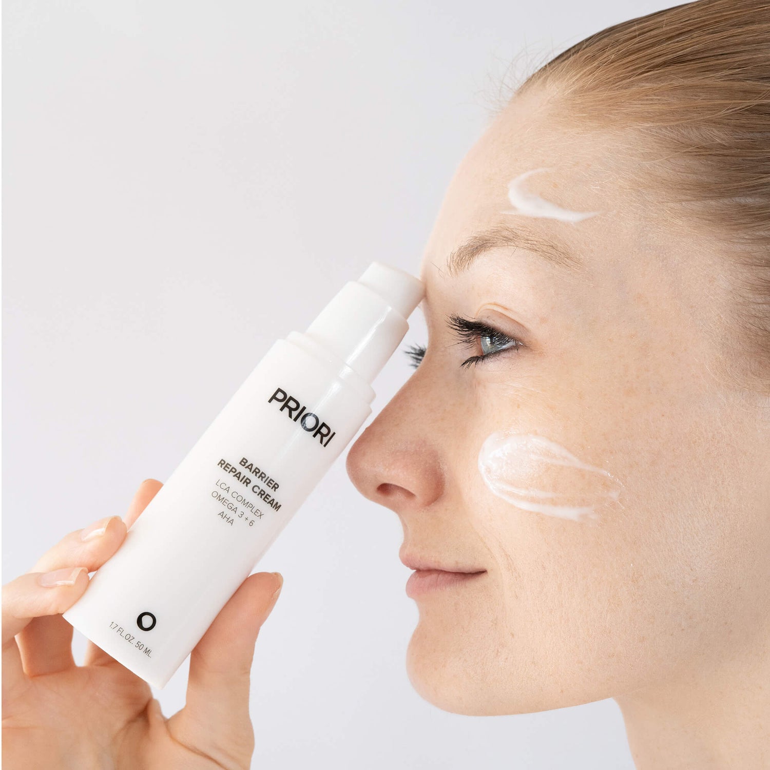 A woman applies PRIORI Skin Barrier Repair Cream to her face while holding the white pump bottle in her hand against a clean white background.