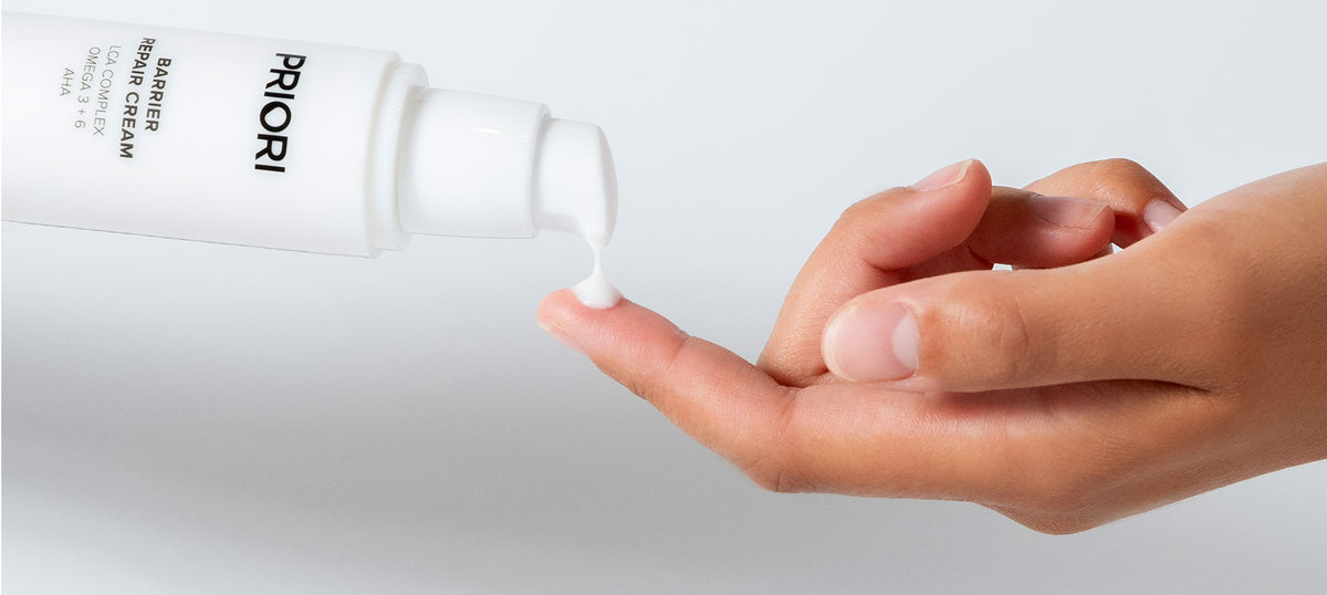 A close-up of a hand receiving a small dollop of PRIORI Barrier Repair Cream with Omega 3 and 6 vitamins from a white pump bottle, with the PRIORI brand name partially visible on the tube against a clean white background.