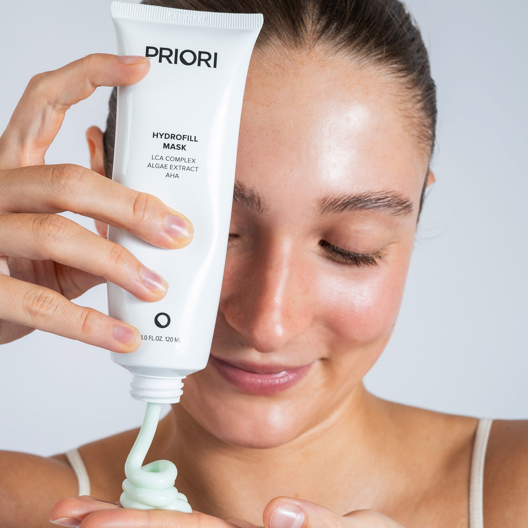 A woman holds a white tube of PRIORI Hydrofill Mask, with pale green gel being squeezed onto her hand against a light gray background.