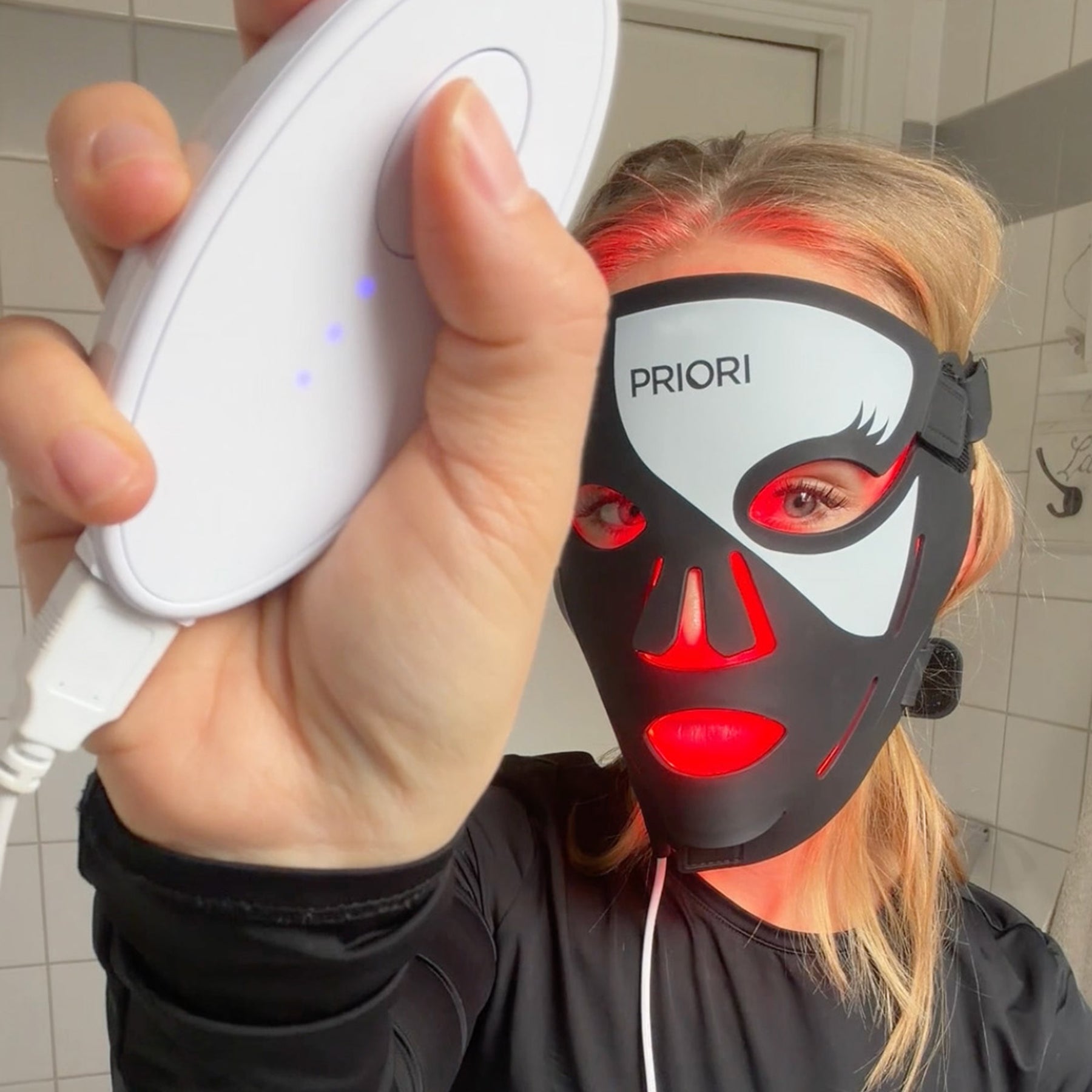 PRIORI Next Generation Red Light Therapy LED Mask with 3 wavelengths for complete skin repair modeled on a blonde woman showing rechargeable mask and controller.