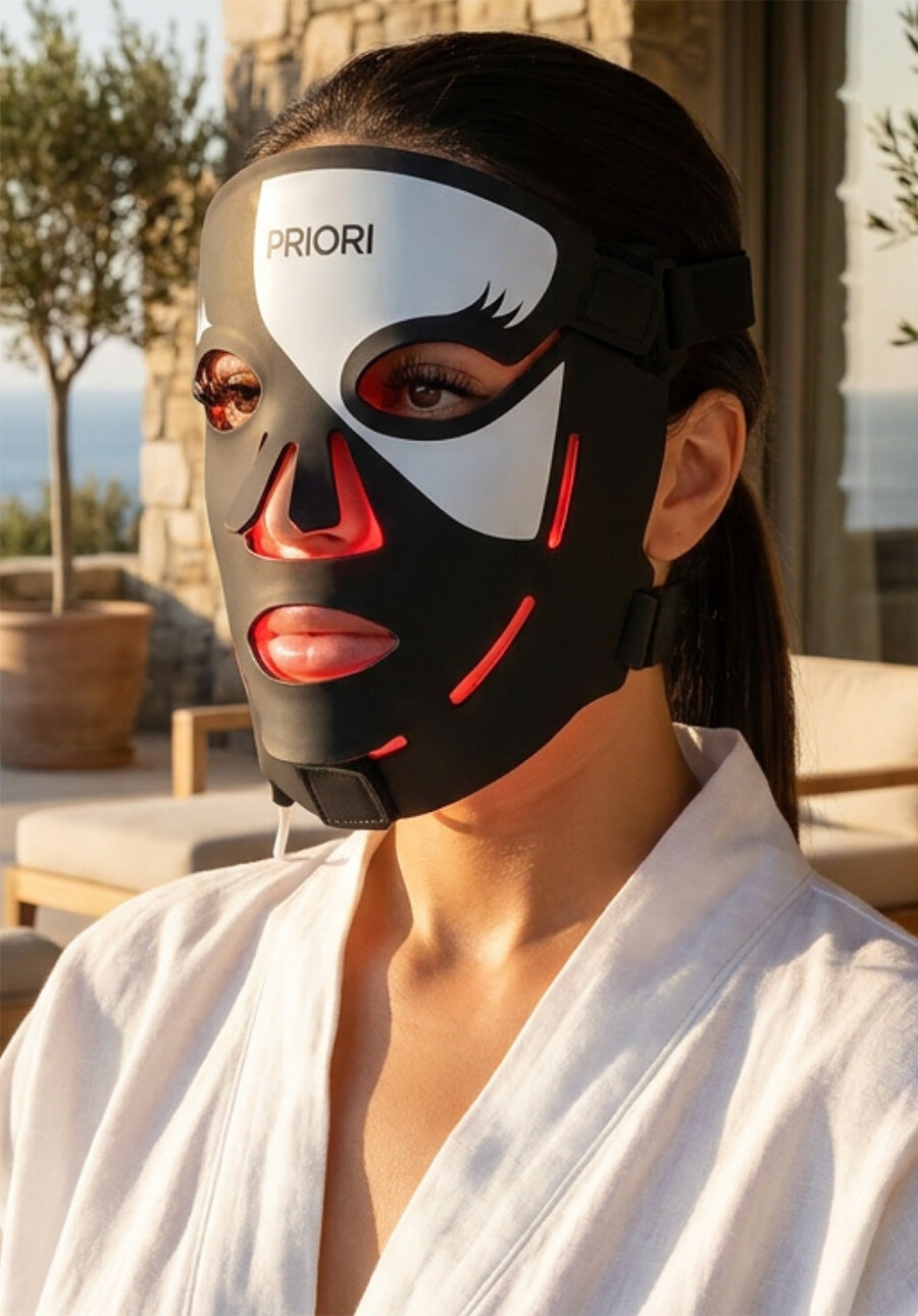 A woman wearing a black, white, and red LED facial mask with the brand name 'PRIORI' printed on it, set against a warm, sunlit outdoor patio background.
