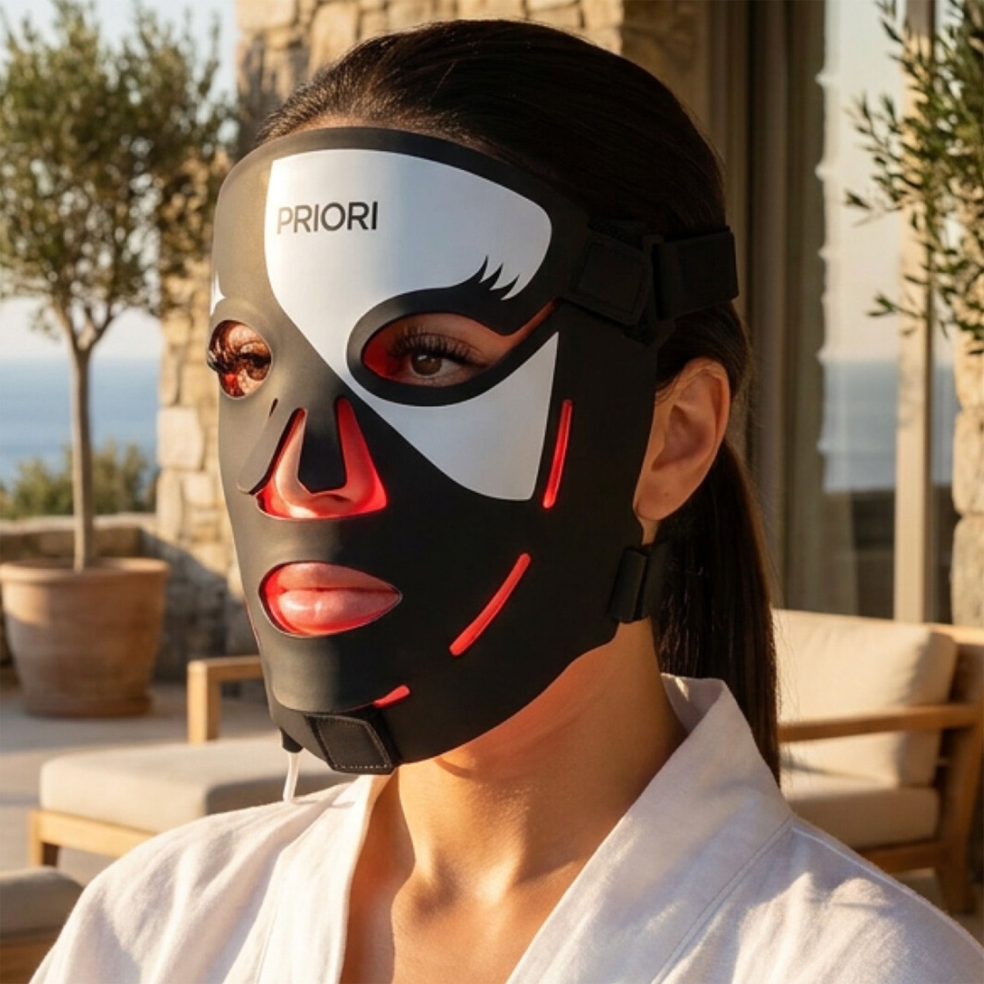 PRIORI Face Red Light Therapy LED Mask in Black and White closeup on model sitting outside at a villa.