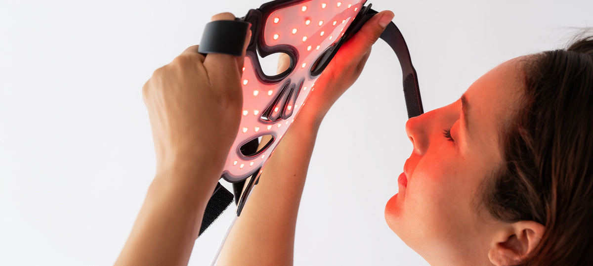 A close-up lifestyle shot of a woman applying or using an anti-aging red light therapy face mask on her face, with the mask glowing pink and red against a clean white background.