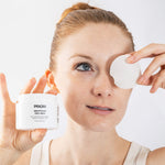 A woman holds a white jar of PRIORI Smoothing Peel Pads next to her face, where she is applying or removing a round white peel pad over one eye. The background is a clean, bright white studio setting.