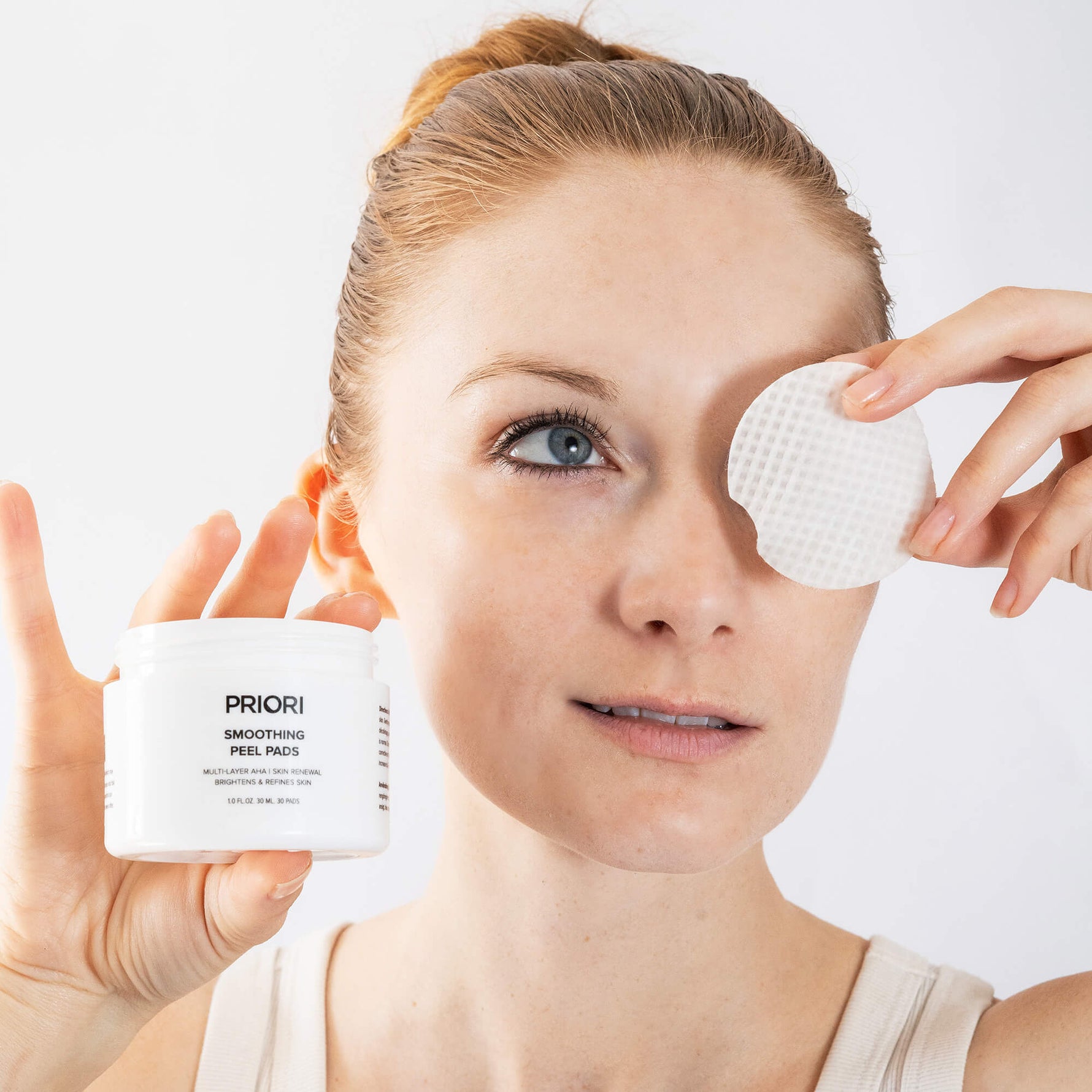 A woman holds a white jar of PRIORI Smoothing Peel Pads next to her face, where she is applying or removing a round white peel pad over one eye. The background is a clean, bright white studio setting.