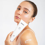 A woman holding a white tube of PRIORI Face Peel & Scrub Deep Cleanser, with exfoliating scrub visible on her cheek and shoulder, against a clean white background.