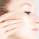 Close-up of a woman applying PRIORI Age Renew Retinol Resurfacing Moisturizing Cream to her cheek with her finger, showing a clean, pale face against a light gray background.