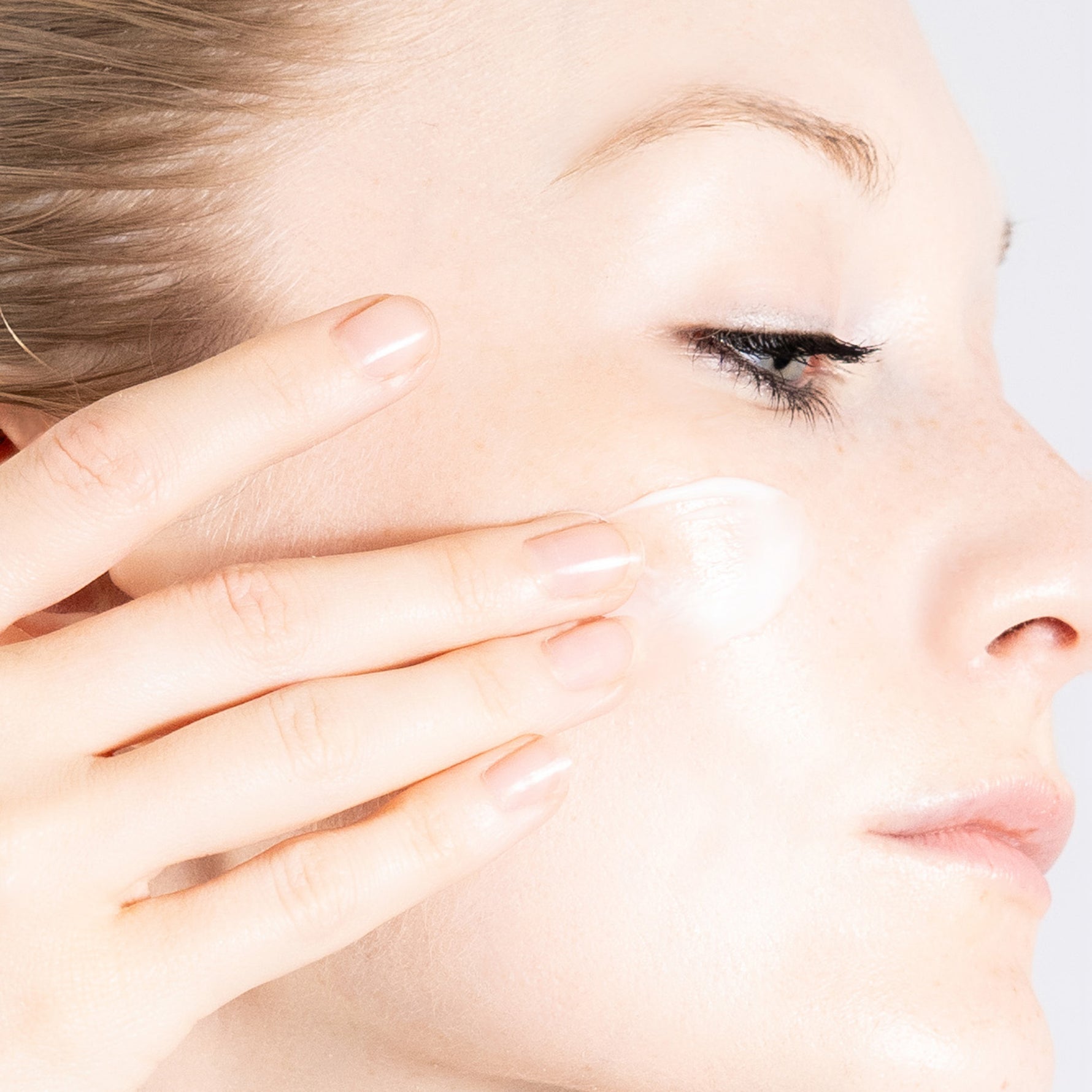 Close-up of a woman applying PRIORI Age Renew Retinol Resurfacing Moisturizing Cream to her cheek with her finger, showing a clean, pale face against a light gray background.