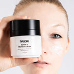 A close-up of a woman holding a white and black jar of PRIORI Repair & Protect Moisturizer Cream with hyaluronic acid next to her cheek, which has a smear of the white cream.