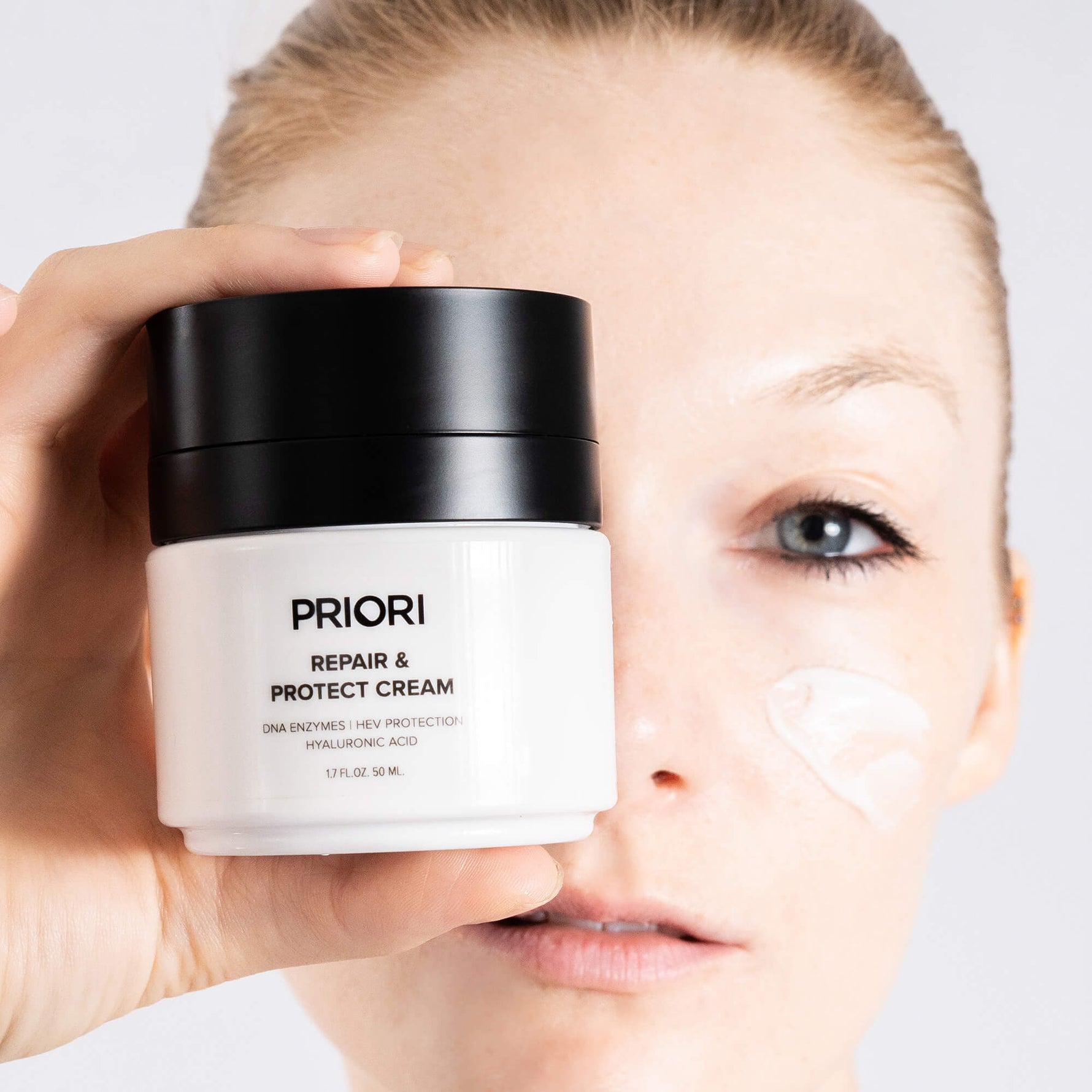 A close-up of a woman holding a white and black jar of PRIORI Repair & Protect Moisturizer Cream with hyaluronic acid next to her cheek, which has a smear of the white cream.