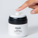 A close-up of a hand pressing the lid to dispense a dollop of daily moisturizer from a white and black PRIORI Repair & Protect Cream pump jar against a light gray background.