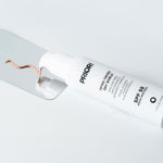 A white tube of PRIORI Tetra Tinted Sun Shield SPF 55 sunscreen lies diagonally on a light gray reflective surface, with a copper-colored smear of product near the cap.