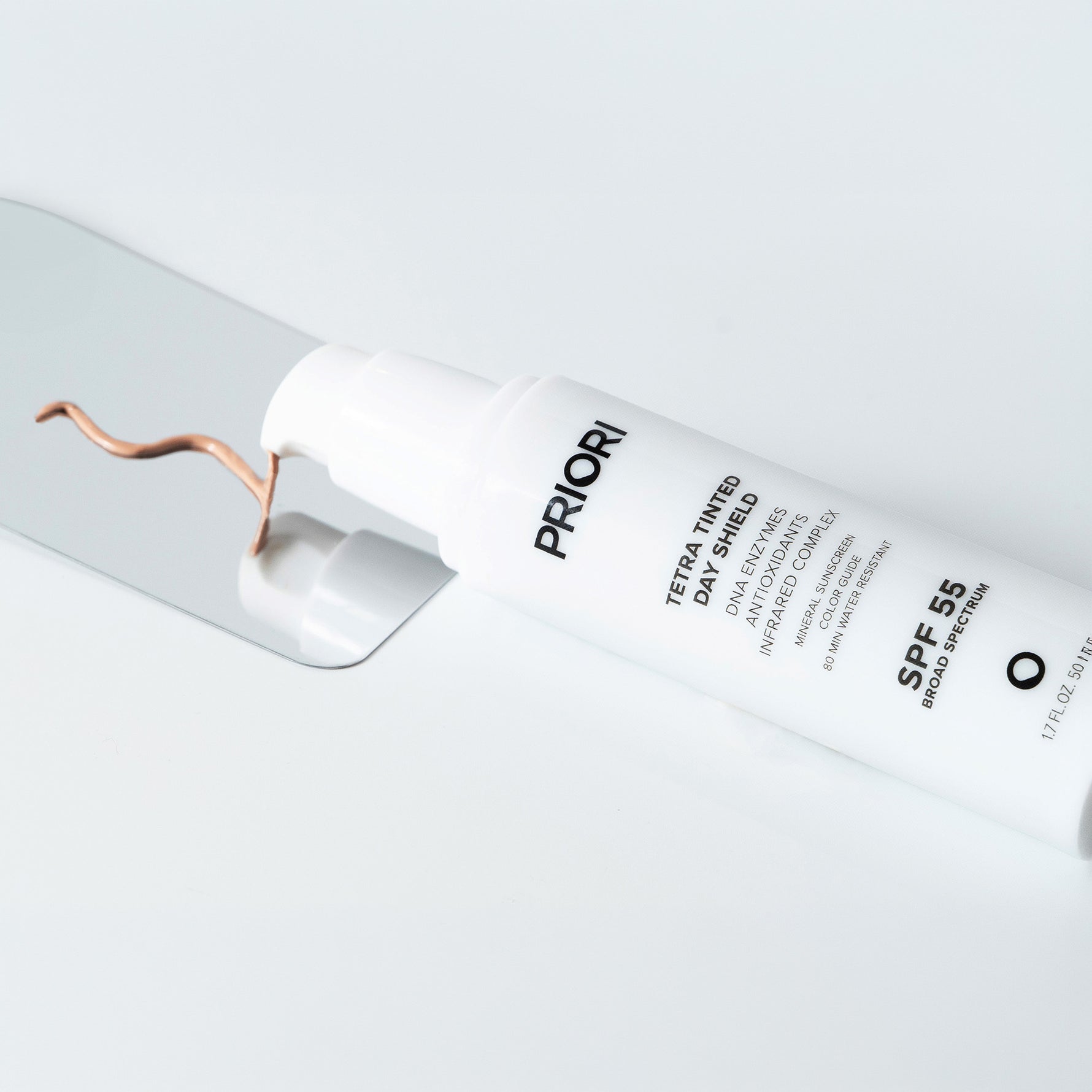 A white tube of PRIORI Tetra Tinted Sun Shield SPF 55 sunscreen lies diagonally on a light gray reflective surface, with a copper-colored smear of product near the cap.