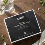 PRIORI Try Me Sample Kit of Cleanser, Serum, and Barrier Restore Moisturizer in travel size tubes in a gift set box on a wooden table with flowers